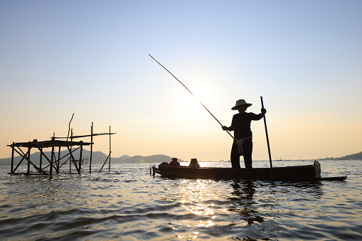 Fisherman casting his net on during sunrise.Silhouette Asian fisherman on wooden boat casting a net for freshwater fish,Silhouette Fisherman Fishing Nets on the boat.Thailand