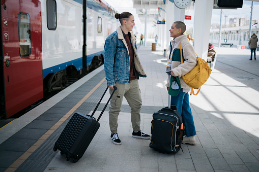 Two young women tourists with luggage standing on train station and talking while waiting for their train. Female friends travellers going on vacation waiting for their train on railway platform with luggage.