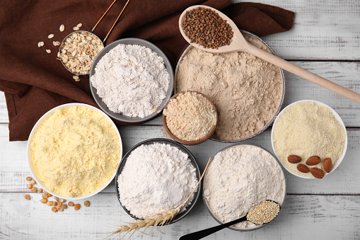 Bowls with different types of flour and ingredients on white wooden table, flat lay