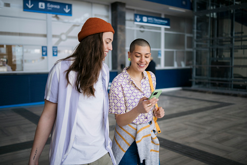 Female couple commuters using mobile phone and smiling on train station. Girlfriends looking at cell phone while waiting for train at railway station.