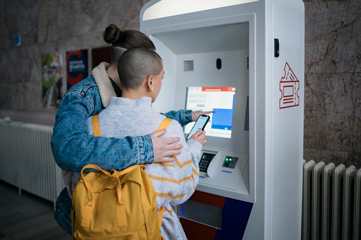 Female traveler couple holding mobile phone buying a train ticket at the automatic vending machine on platform. Gay couple booking train ticket and paying with digital wallet at automatic ticket machine at railway station.
