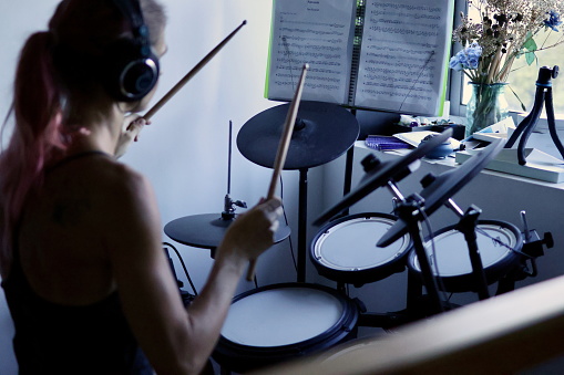 A young woman wearing headphones plays the drums in front of the window