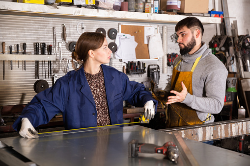 Focused bearded foreman talking to young female apprentice measuring metal structures in metalworking workshop