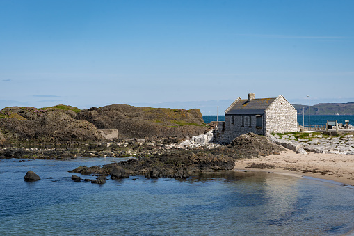 Small stone house on the coast at Ballintoy harbour, County Antrim, Northern Ireland, on a sunny summer day with blue sky. It has a rugged rocky coastline and is a popular tourist attraction.