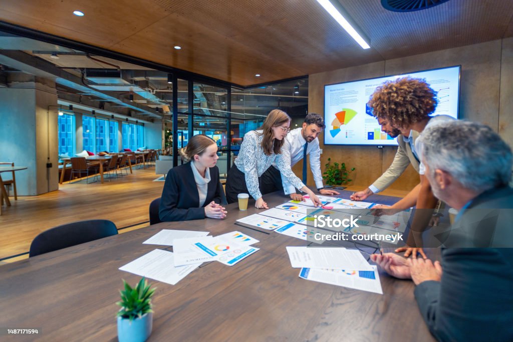 Paperwork and group of peoples hands on a board room table at a business presentation or seminar. Paperwork and group of peoples hands on a board room table at a business presentation or seminar. The documents have financial or marketing figures, graphs and charts on them. People are pointing at the data. There is a T.V. with financial data in the background Business Stock Photo