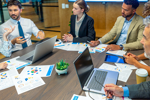 Diverse group of people meeting and working at a board room table at a business presentation or seminar. The documents on the conference table have financial or marketing figures, graphs and charts on them.