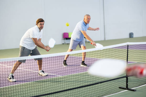 uomo adulto concentrato che gioca la partita di pickleball in squadra con il partner più anziano - tennis men indoors serving foto e immagini stock