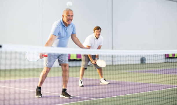 dois homens atléticos de idades diferentes est�ão jogando uma partida de pickleball em uma quadra dentro de uma instalação esportiva - tennis men indoors playing - fotografias e filmes do acervo