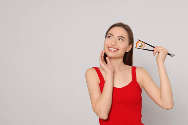 Happy young woman holding sushi roll with chopsticks on light background. Space for text
