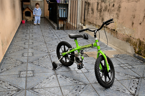Baby boy playing in the backyard of his house. It is possible to see that the child is looking at the bicycle that is in the foreground of the scene. The photo was taken outside the residence.