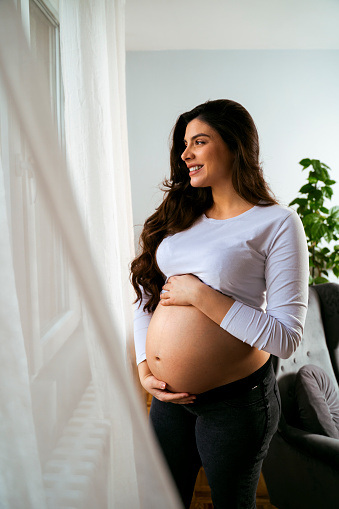 Portrait of Young beautiful pregnant woman holding her belly and smiling, looking through window