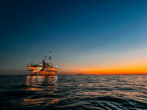 A stunning photograph of offshore oil drilling at sunset in Huntington Beach, California. The pink hues of the setting sun highlight the industrial machinery and equipment used in the drilling and extraction of fossil fuels, including crude oil and natural gas. 

This image captures the intersection of the energy industry and the natural beauty of the Pacific Ocean, and speaks to issues of fuel and power generation, energy crises, and environmental concerns surrounding the oil and gas industry.