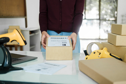 Close up of young female online business, store owner or entrepreneur standing at desk with laptop, scanner, and cardboard boxes holding and putting label tag on parcel for preparation before shipment, delivery to customer. Online business, e-commerce, logistics concept.