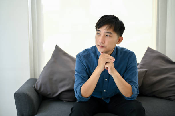 istock Thoughtful Asian man sits on a sofa, pondering and looking out the window. 1487220455