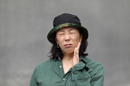 Asian woman with hat and hand to face with an uncomfortable expression. Gray background.