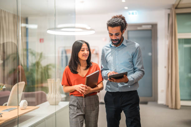 Diverse Colleagues Working Together on Digital Tablet Two handsome, diverse colleagues are seen engaging in a productive conversation while utilizing wireless technology and a digital tablet in a modern office setting. Their smart casual attire adds to their professional appearance as successful employees. business casual stock pictures, royalty-free photos & images