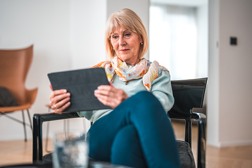 Attractive senior White female sitting at home, using a digital tablet to read an e-book. She is wearing jeans and casual clothes.