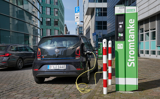 Düsseldorf, Germany - April 27, 2023: A black Volkswagen „e-up!“ in front of Media Harbor office buildings connected to a recharging station. The green recharging station is from the local energy provider of the city Düsseldorf. Focus on the car.