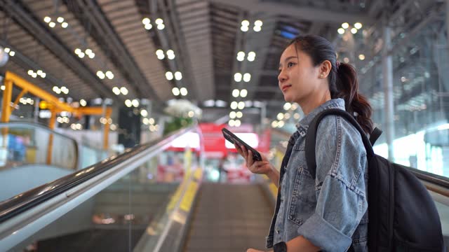 Asian female traveler using a smartphone in an airport. She is using an escalator.