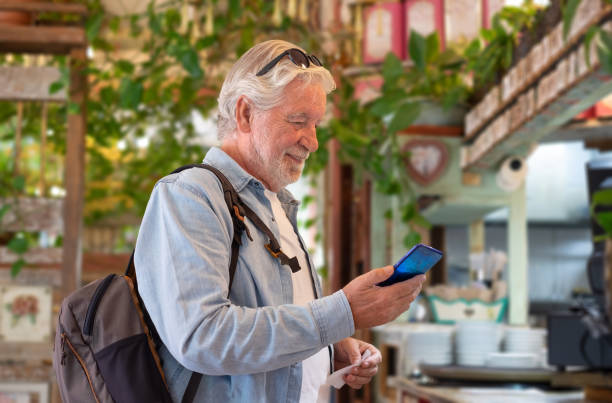Smiling senior white-haired man pays at the coffee shop checkout using the application on his mobile phone. Concept of modernity and online electronic payments