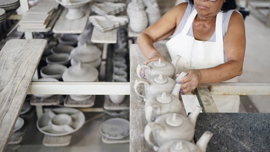 Cropped shot of a female worker arranging handmade clay teapots for drying on rack at ceramic manufacturing factory