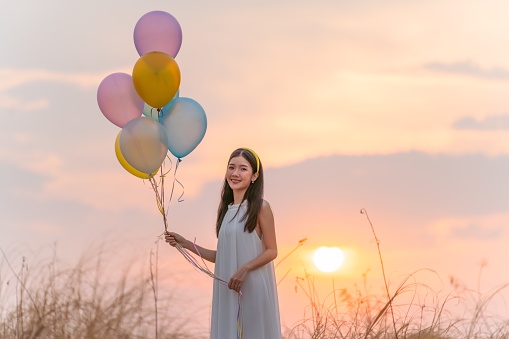 Portrait photo of a young beautiful asian woman smile happily holding a bunch of balloons at an outdoor wide open grass field during sunset moment