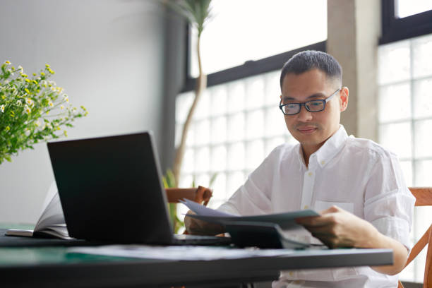 istock An Asian man sits at his dining table, poring over a project document and working on his laptop, illustrating the concept of remote work from home 1487248294