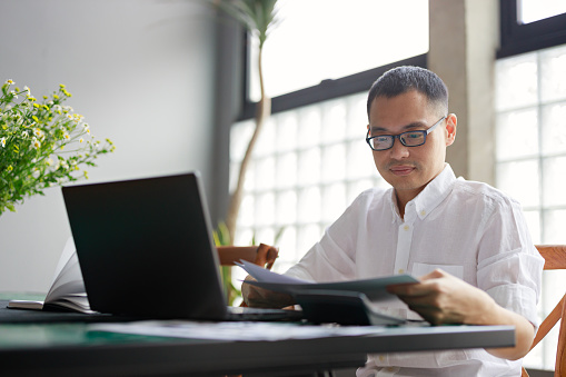 An Asian man sits at his dining table, poring over a project document and working on his laptop, illustrating the concept of remote work from home