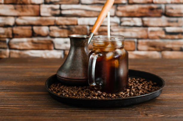 Iced coffee in transparent glass and roasted coffee beans Glass transparent mug with iced coffee and cezve,coffee grains, against the background of a stone wall and a wooden table turkish coffee pot cezve stock pictures, royalty-free photos & images