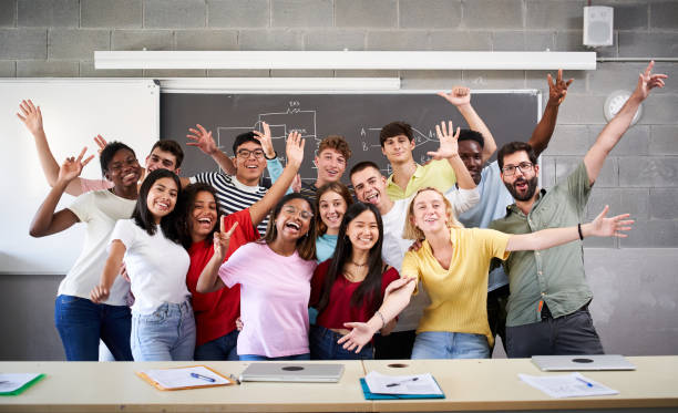 Portrait of a cheerful group of students celebrating in class looking at the camera. Happy Young college people of different ethnicities posing for a photo in the classroom hands up stock photo
