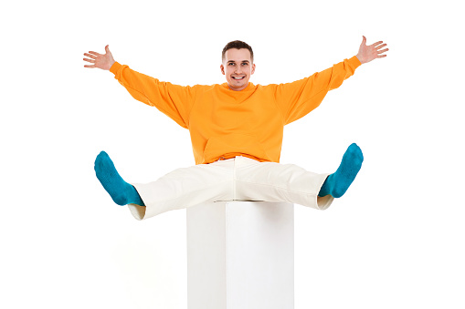 young hipster student man in orange sweatshirt have fun sitting on cube on white background. Full length