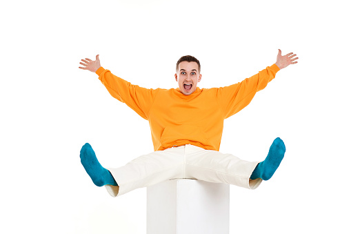 young funny man in orange sweatshirt sitting on white cube on white background. Full length