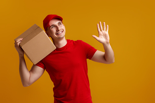 happy delivery man employee in red cap, red t-shirt holding cardboard box isolated on yellow background