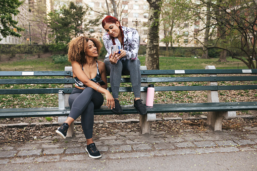 Two friends in their morning activity at the park, active lifestyle in Spring. They are resting on park bench after a stressful hard training class. Playing on the phone.
