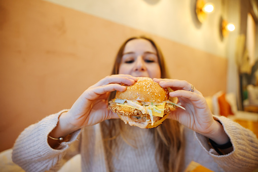 Happy healthy woman sitting in indoors food court and eating an delicious hamburger, modern meal concept