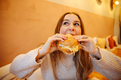 Happy healthy woman sitting in indoors food court and eating an delicious hamburger, modern meal concept