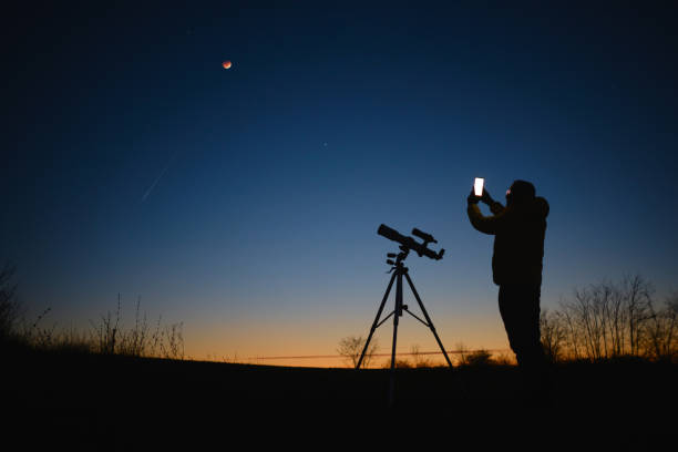istock Astronomer looking at the starry skies with planets, falling stars and Moon eclipse with a telescope while using modern smartphone. 1487260948