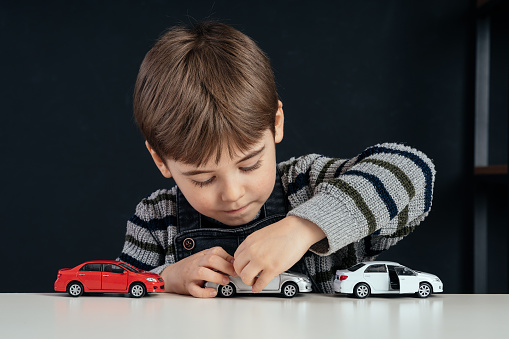 Hands of little child boy playing with multicolored toy cars on white wooden background.