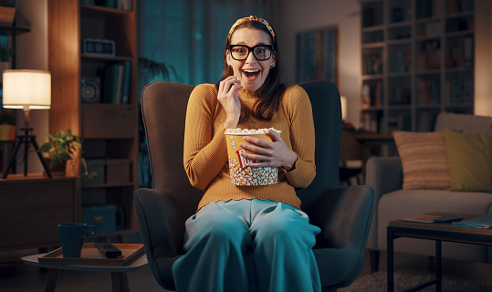 Woman sitting on the armchair at home and watching a comedy movie, she is eating popcorn and smiling