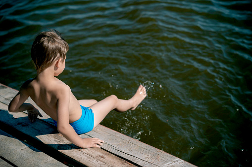 cute caucasian boy sitting on wooden pier diving into lake in countryside