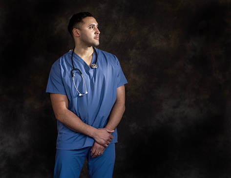 Studio image of doctor on dark backdrop wearing medical scrubs and stethoscope lit with a spotlight. He is looking away from the camera with a contented look on his face.
