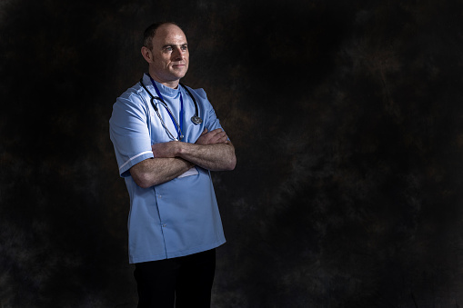 An image of a white doctor standing against a dark background  with his arms folded, is looking off to his left. He is dressed in a  blue uniform top and has a stethoscope and lanyard around his neck.