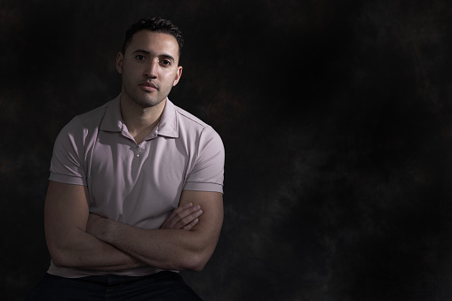 A young man sits against a dark background. He is wearing a short sleeve shirt and his arms are folded, he is looking directly at the camera with a neutral expression.