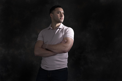A young man stands against a dark background. He is wearing a short sleeve shirt and his arms are folded, he is looking out to his left with a neutral expression.