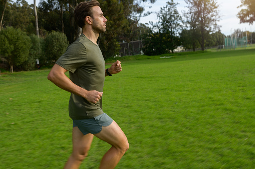 Male runner running on a grass field wearing a green khaki coloured shirt and running shorts.  There are Blue Gum Eucalyptus Trees behind.  Stellenbosch, South Africa.