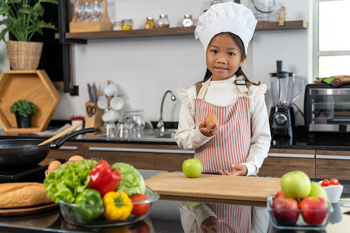 Portrait Asian lovely girl wearing chef hat and apron standing with an egg in hand near counter, bell pepper, vegetable and cooking ware place on top, smile and looking at camera, copy space