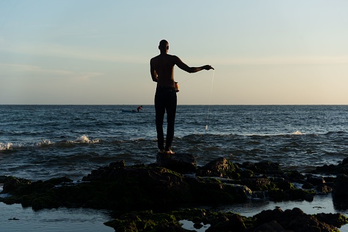 Salvador, Brazil – December 12, 2021: A fisherman on top of the rocks fishing. Late afternoon at Rio Vermelho beach in Salvador, Bahia.