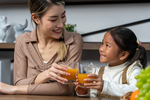 Beautiful mother and her lovely daughter drink fresh orance juice on table in kitchen at house, they talking and smile with happy together, copy space