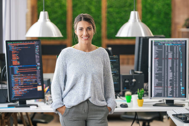 Retrato de jovem programadora sorridente, sentada em sua mesa com computadores em um escritório de TI - foto de acervo