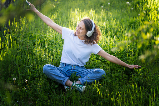 Young cheerful dreamy woman listening to music using smartphone and headphones sitting in summer garden,